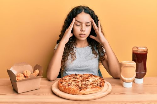 Menina hispânica adolescente comendo pizza e frango frito com a mão na cabeça, dor de cabeça por causa da alimentação, sofrendo de enxaqueca.